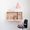 Minimalist home office setup featuring a wall-mounted wooden desk, pink pendant light, and a black chair with stacked books, reflecting modern design aesthetics.