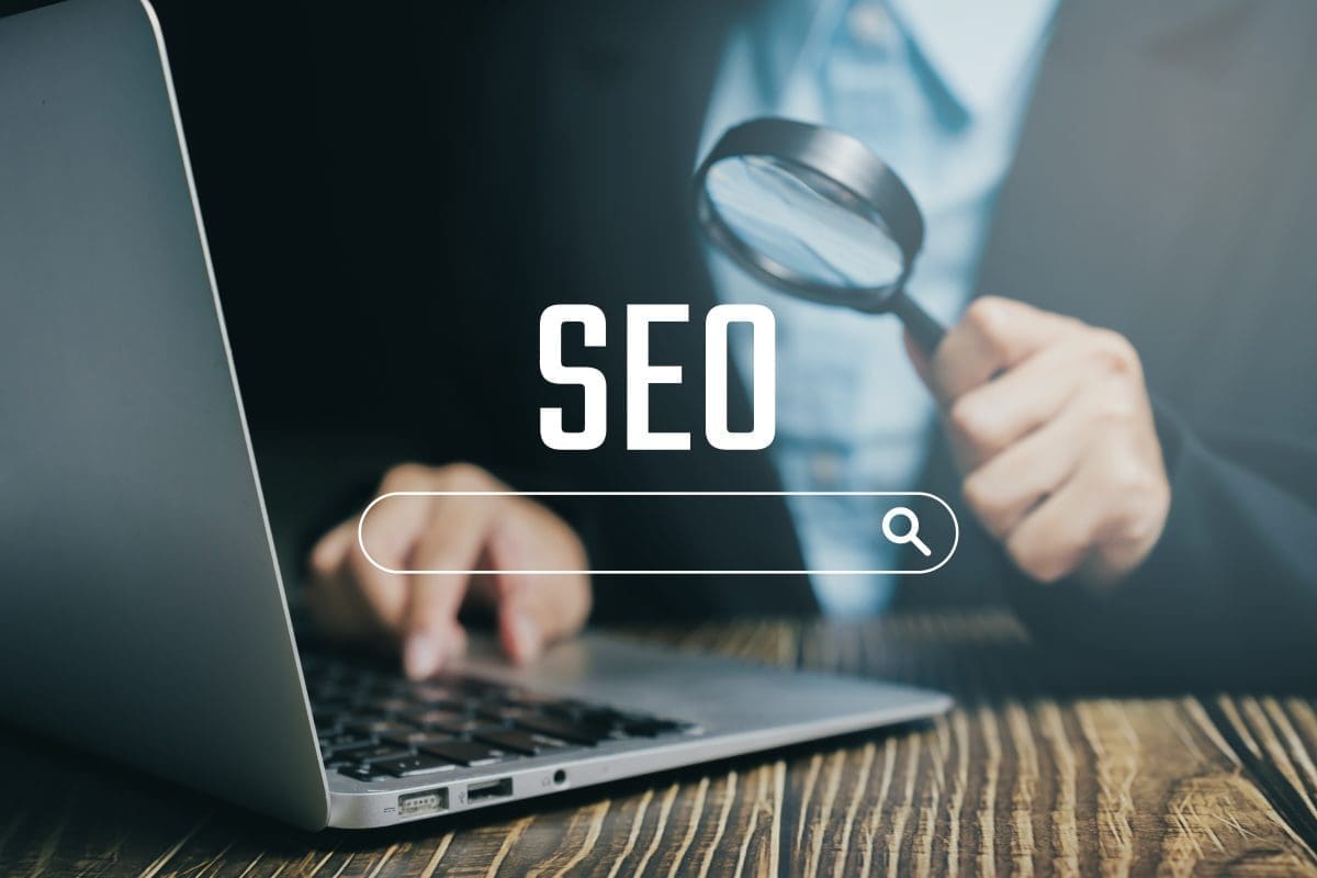 Person using a laptop with a magnifying glass, focusing on SEO strategies and optimization for improved online visibility.